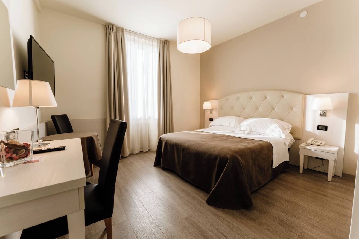 Habitación de hotel con cama, mesitas de noche, escritorio, silla, lámpara y ventana con cortinas.