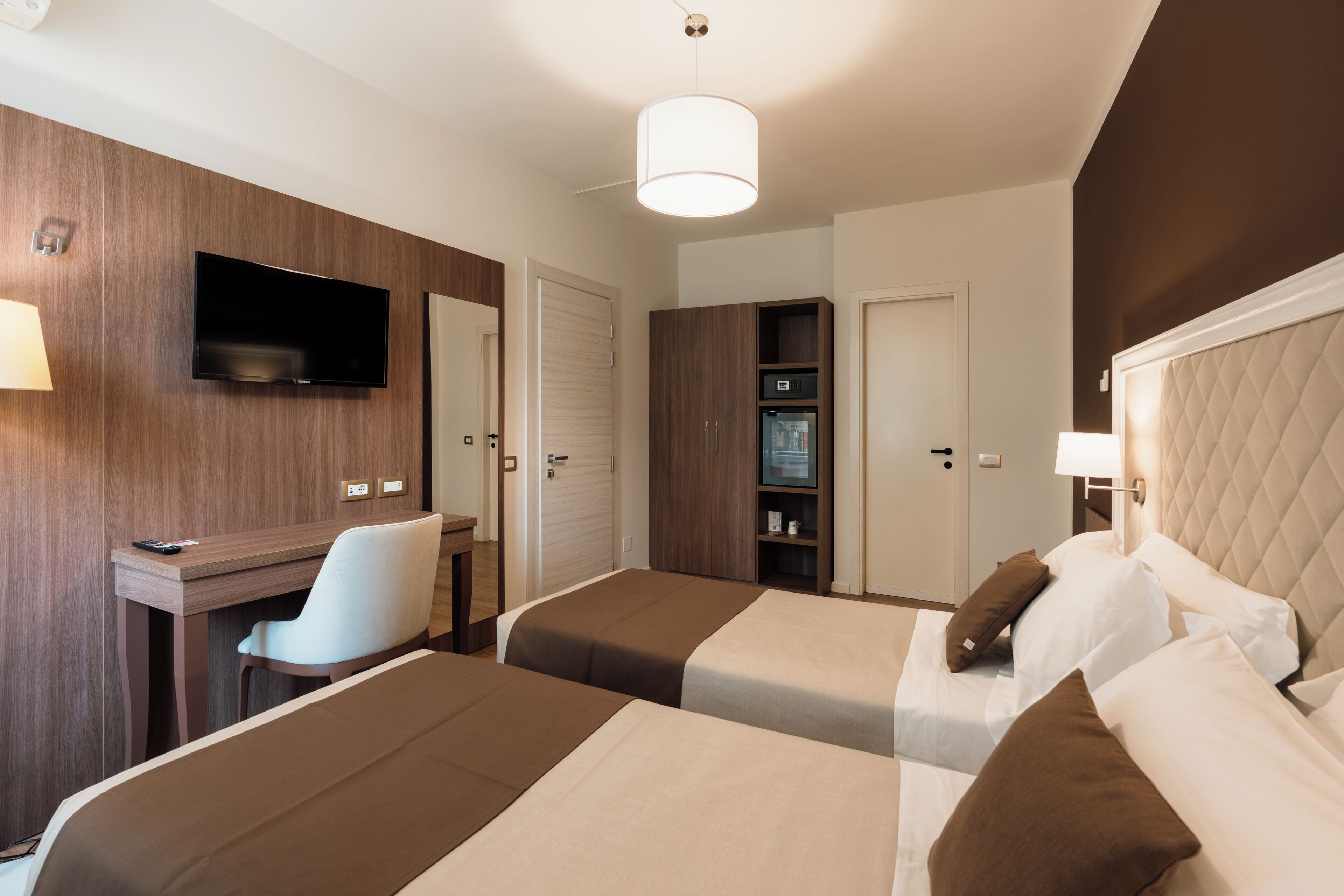 A hotel room with two beds, a desk, a chair, a TV, a wardrobe, and a lamp.