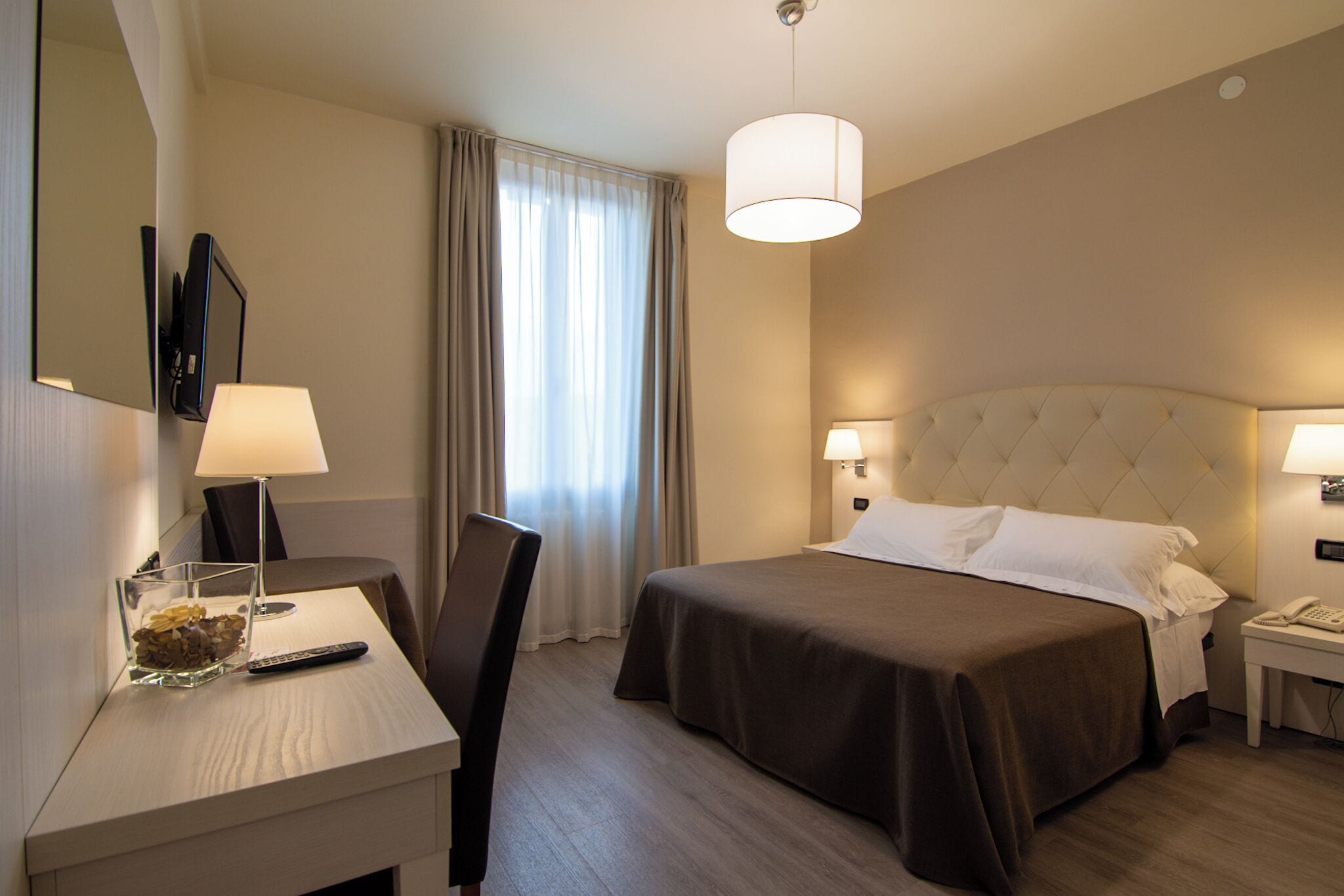 Een hotelkamer met een bed, nachtkastjes, een bureau met lamp, een stoel, een spiegel en een raam met gordijnen.
