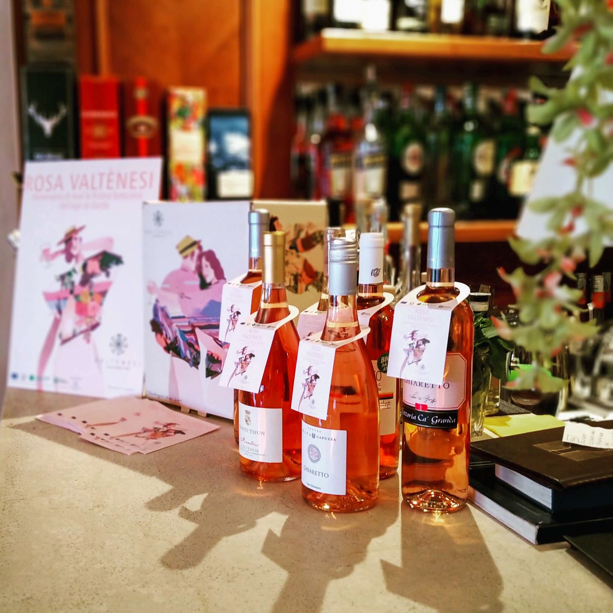 Botellas de vino rosado con etiquetas y adornos, dispuestas sobre una barra con un cartel al fondo.