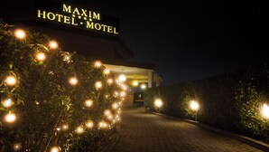 Een nachtzicht van de ingang van MAXIM HOTEL MOTHET, met verlichte borden en een geplaveid pad dat naar de ingang leidt.