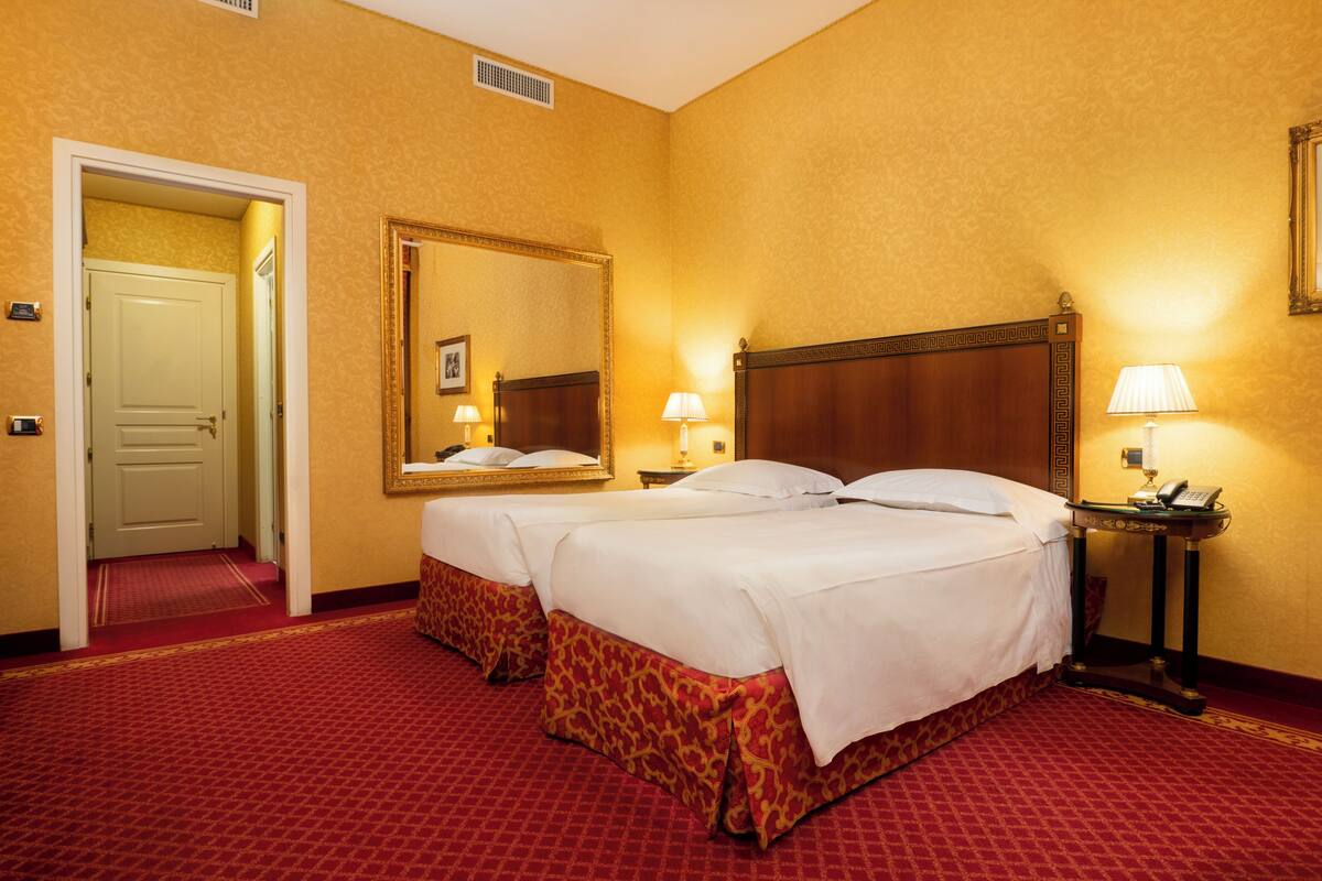 A hotel room with two beds, a large mirror, a bedside table with a lamp, and a door leading to another room.