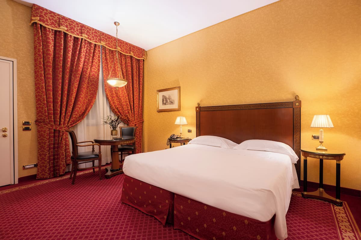 A hotel room with a large bed, two bedside tables with lamps, a desk with a chair, and a door.