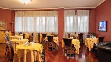 Uma sala de jantar com mesas preparadas para o serviço, uma tela de televisão e uma janela grande com cortinas.