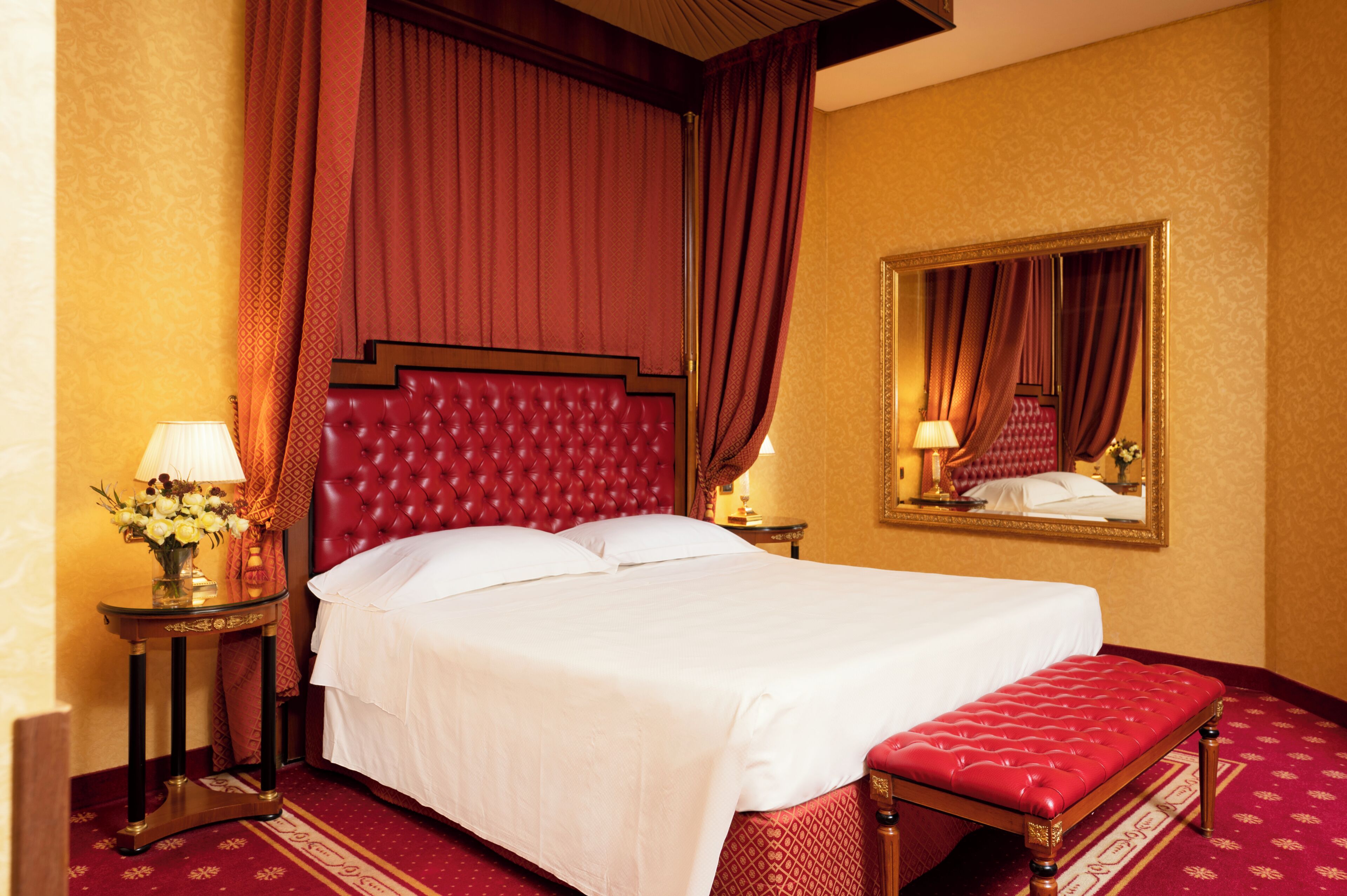 A hotel room with a large bed, a red upholstered bench, a bedside table with a lamp, and a mirror on the wall.
