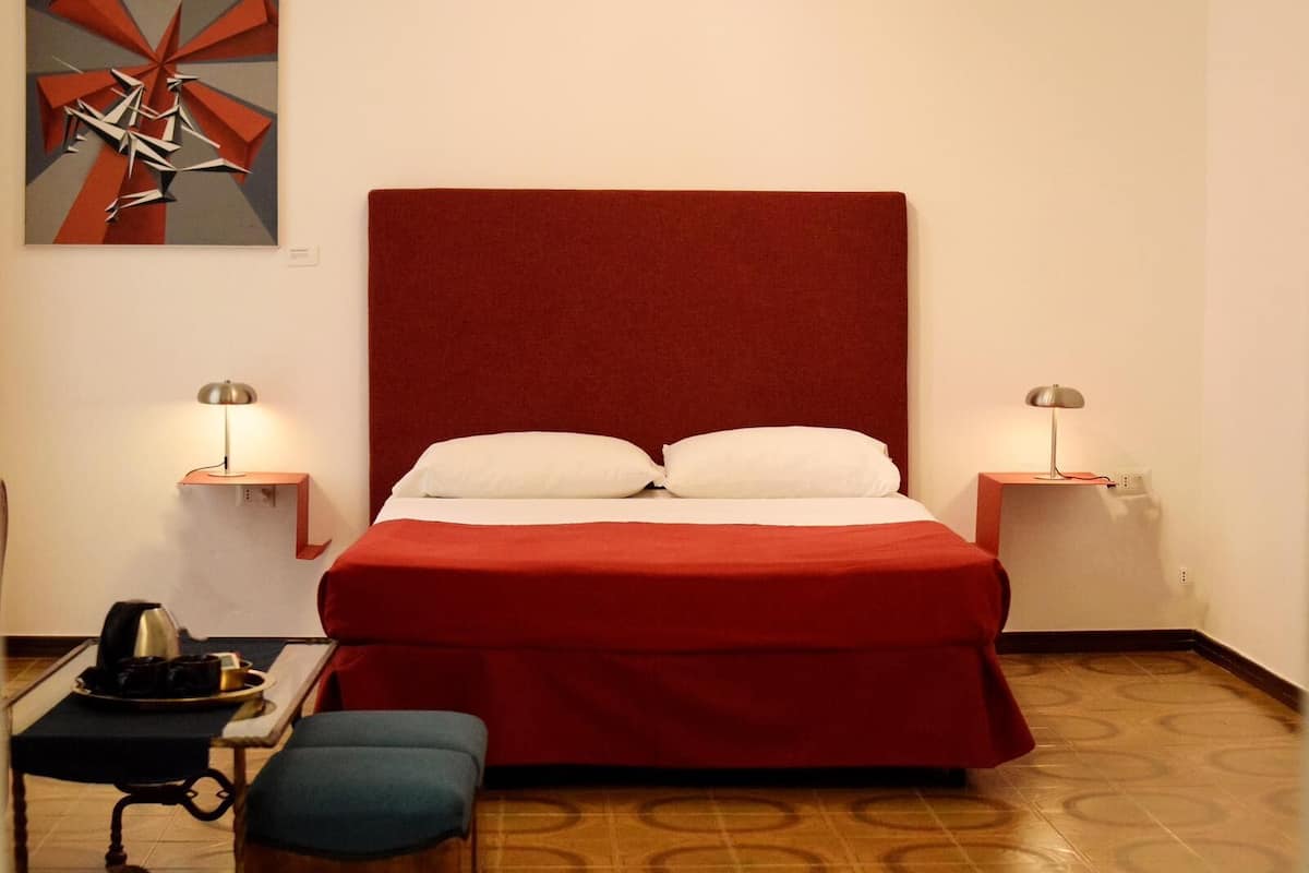 A hotel room with a large bed, two bedside tables, a small table with a tray, a bench, and a patterned floor.