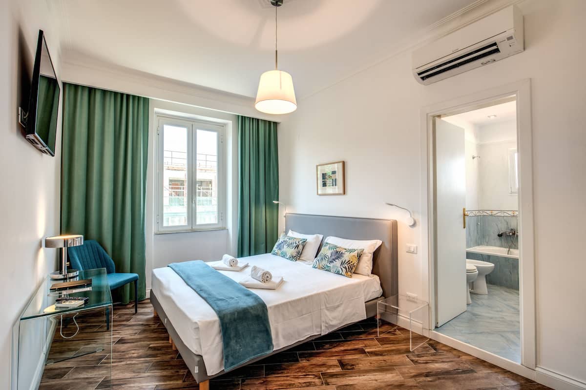 A hotel room with a bed, a desk, a chair, a window with curtains, an air conditioning unit, and a bathroom visible through an open door.
