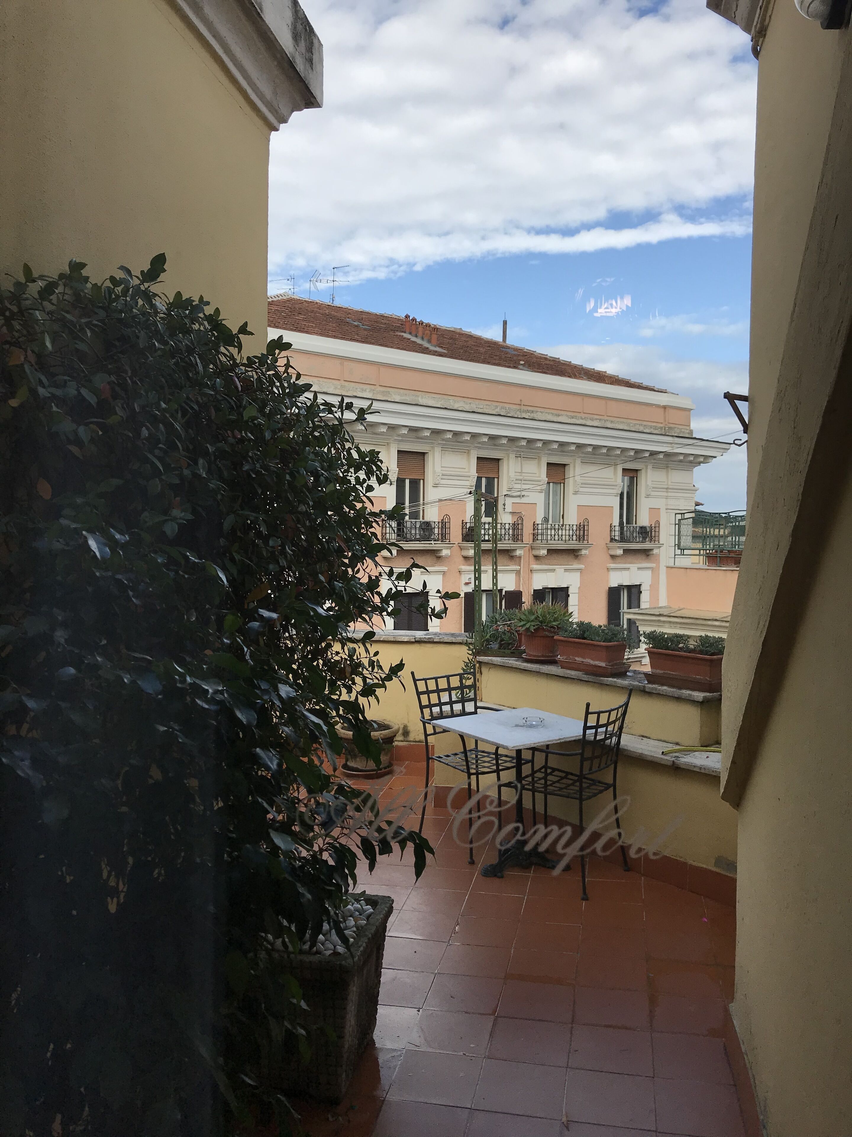 A balcony with a table and chairs, potted plants, and a building in the background.
