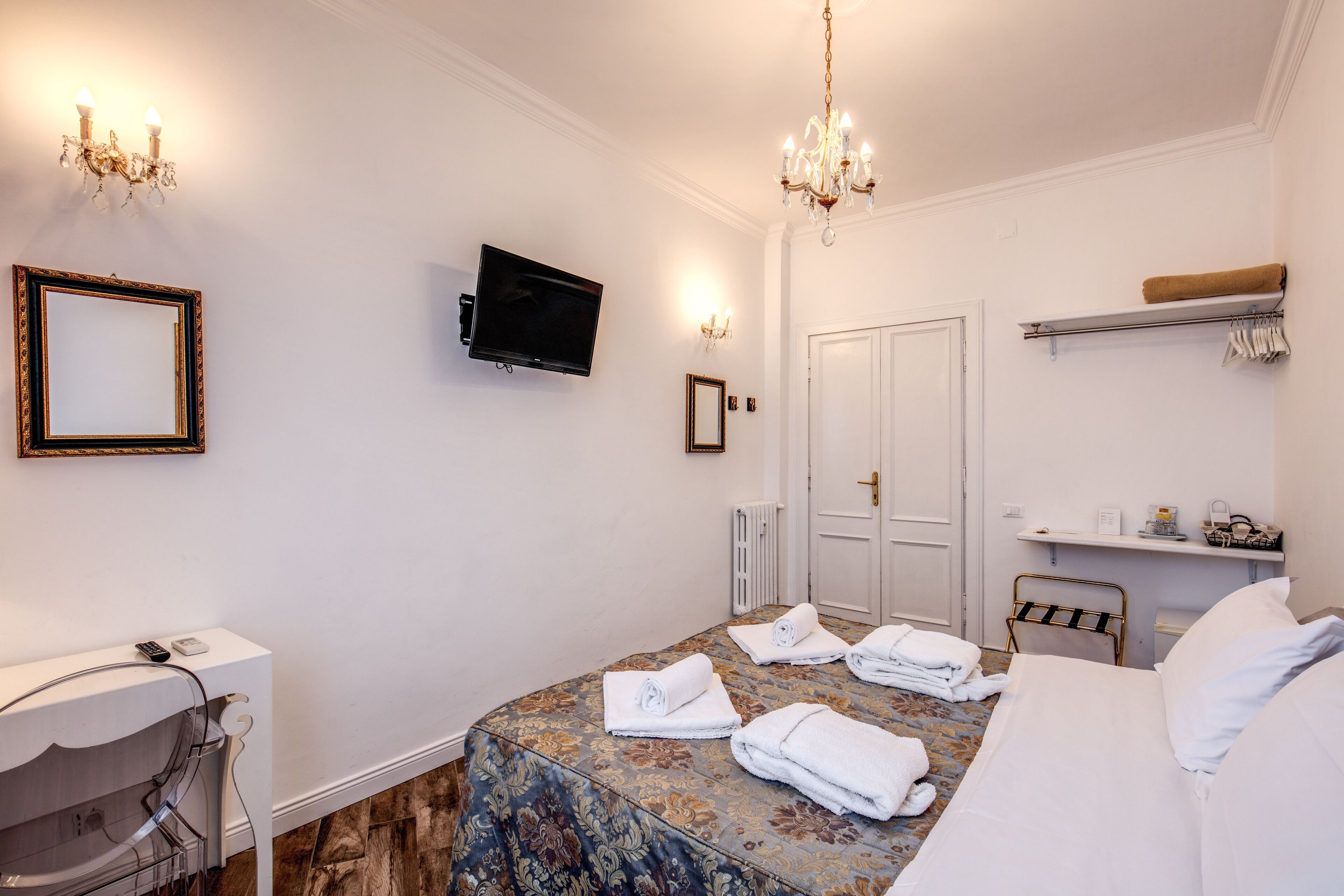 A hotel room with a bed, a television, a mirror, a chandelier, and a bathroom area.