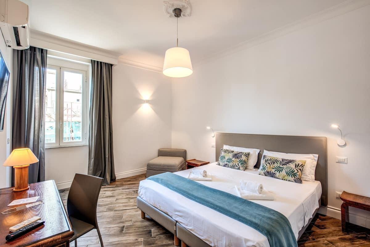A hotel room with a bed, a desk with a lamp, a chair, a small stool, and a nightstand.