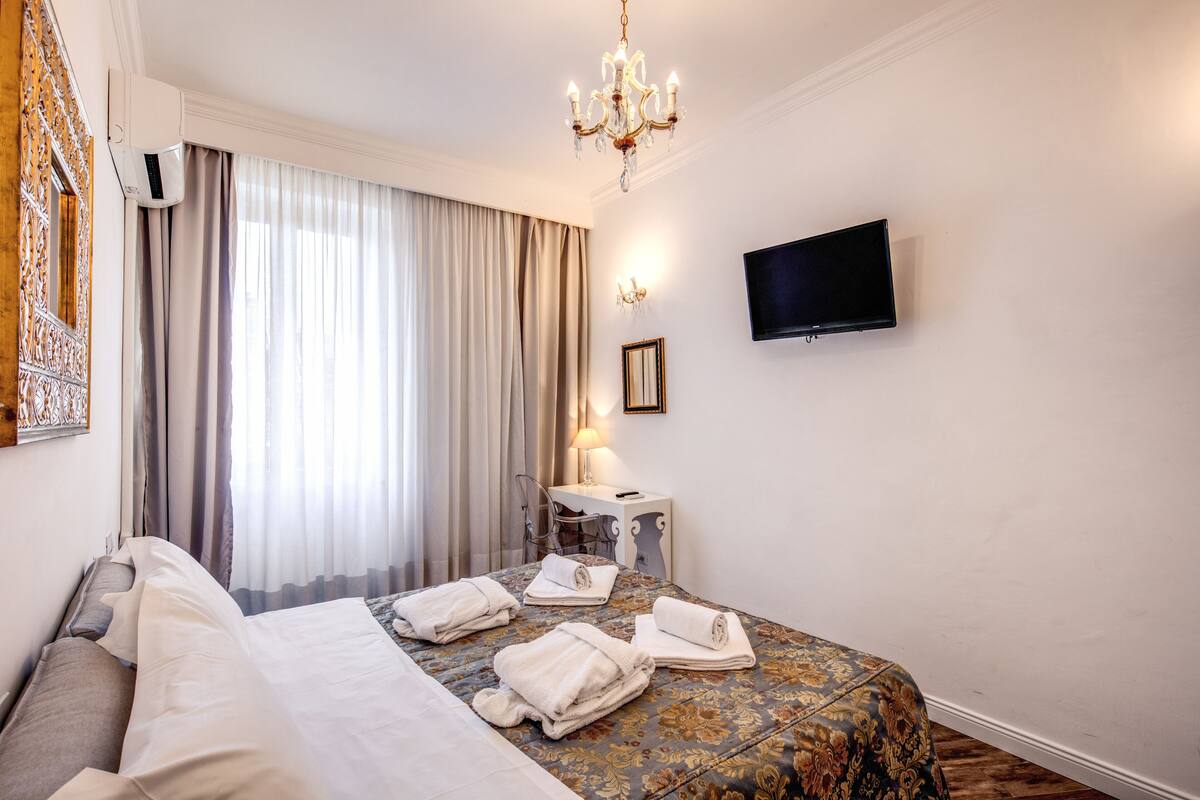 A hotel room with a bed, a desk, a chair, a television, and a chandelier.