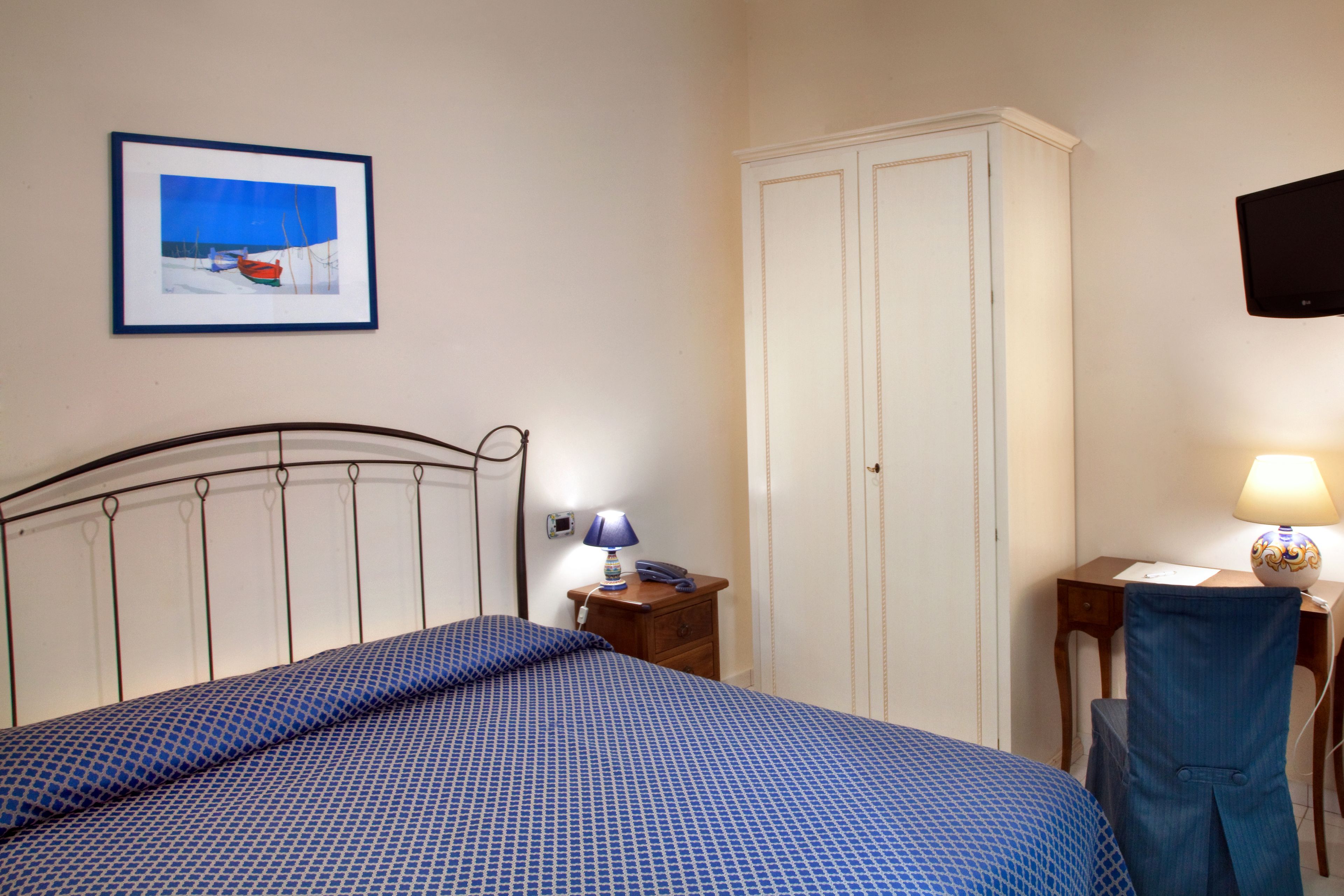 Une chambre à coucher comprenant un lit, une table de chevet, un bureau, une chaise, une armoire, un téléviseur et un tableau encadré accroché au mur.