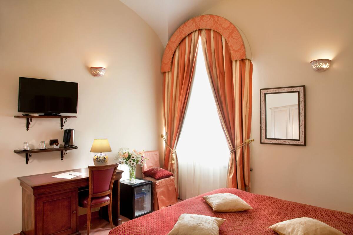 A hotel room with a bed, a desk with a lamp, a TV, and a window with curtains.