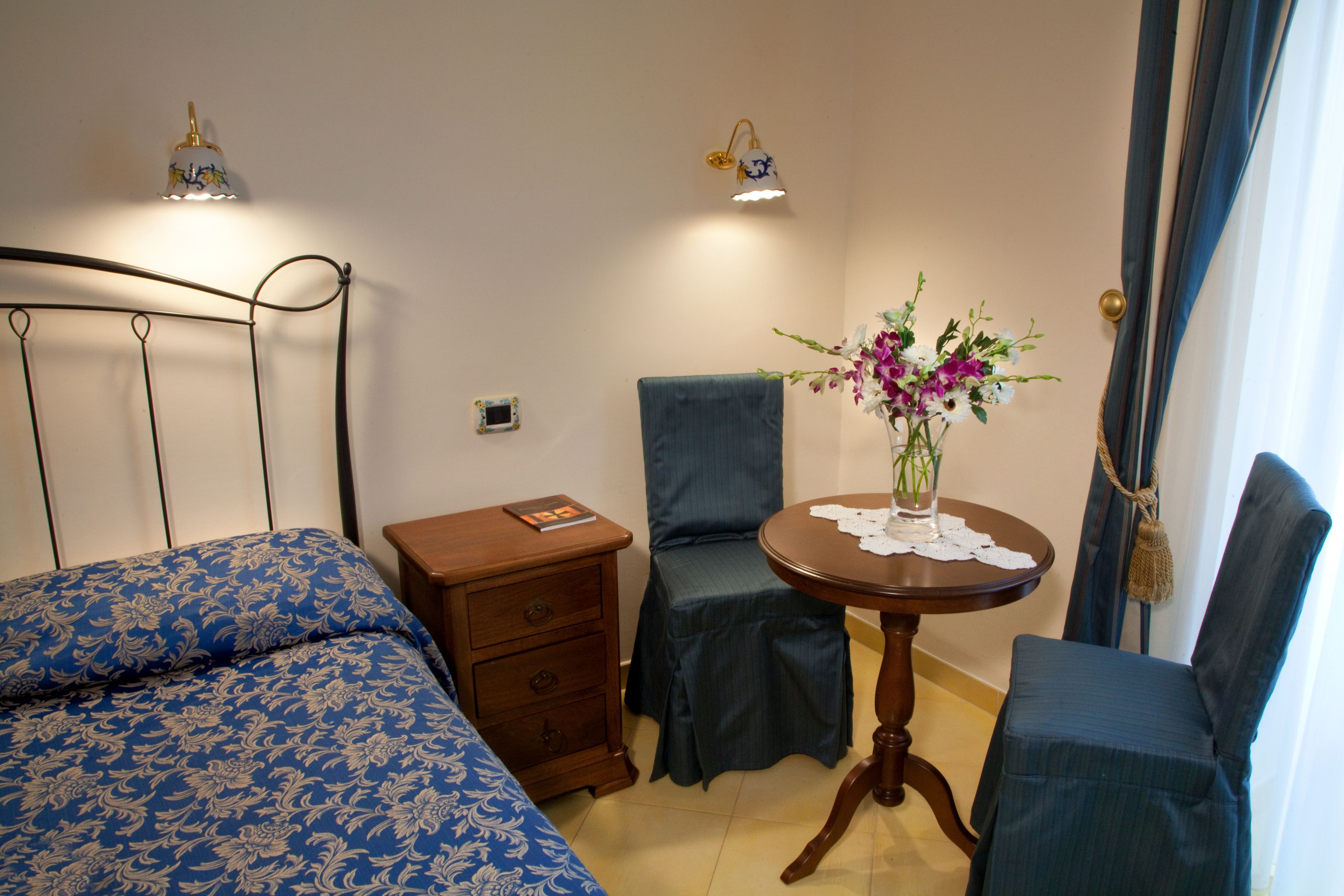 A bedroom with a bed, a nightstand, a chair, a table with a vase of flowers, and two wall-mounted lamps.