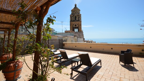 Terraço com cadeiras de descanso, plantas em vasos e vista para uma torre histórica e o mar.