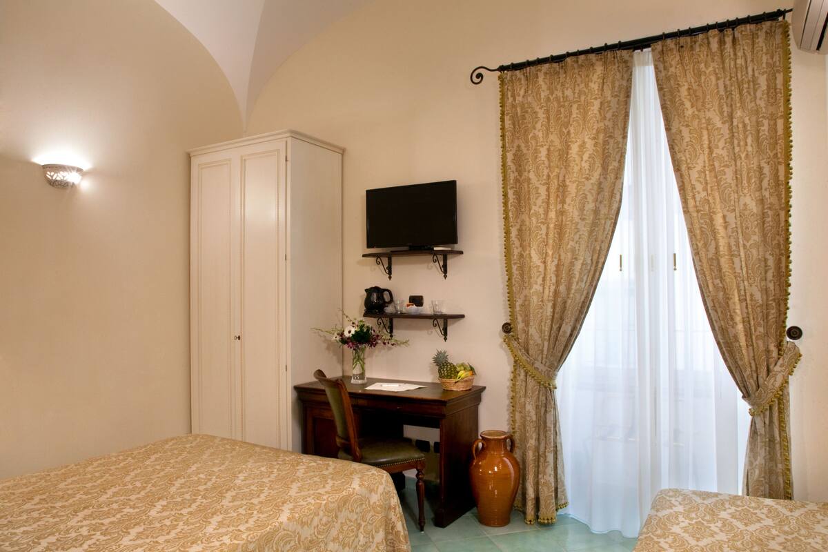 A hotel room with two beds, a desk with a chair, a television, a vase with flowers, and a large window with curtains.