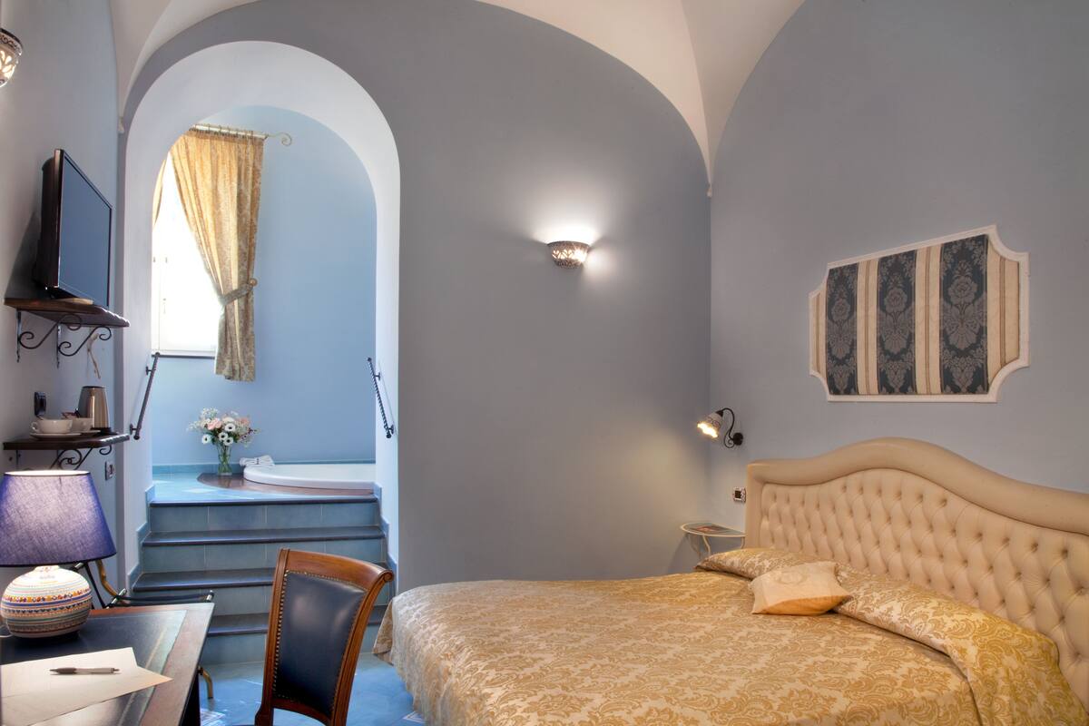 A bedroom with a bed, a desk, a chair, a TV, and a bathroom visible through an archway.