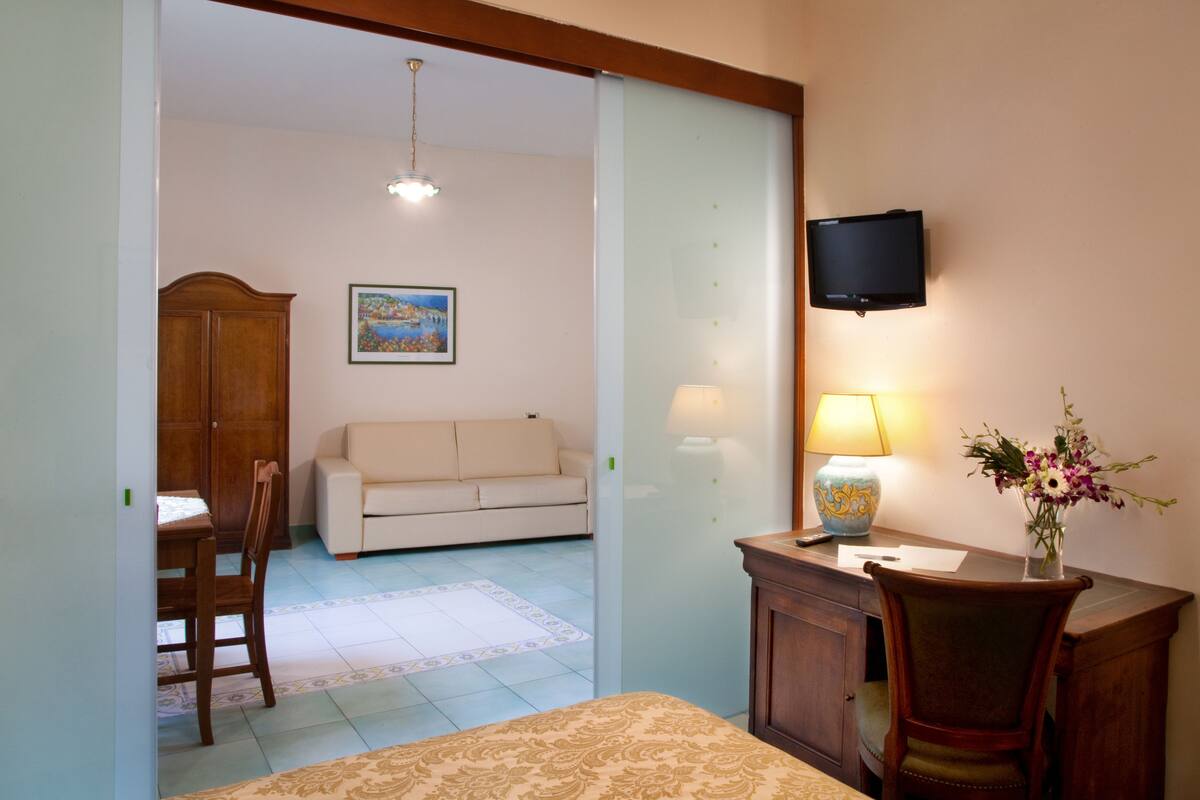 A hotel room with a bed, a desk with a lamp, a TV, a sofa, a dining table with chairs, and a wooden cabinet.