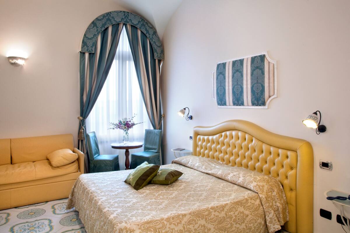 A hotel room with a bed, a sofa, a small table with a vase, and a chair.