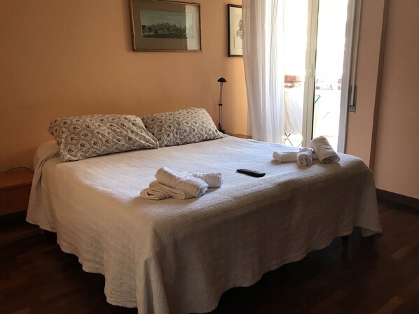 A neatly made bed with floral-patterned pillows, a remote control, and rolled towels on a light-colored bedspread in a room with wooden flooring and a framed picture on the wall. - Le Finestre sul Vaticano (Rome)