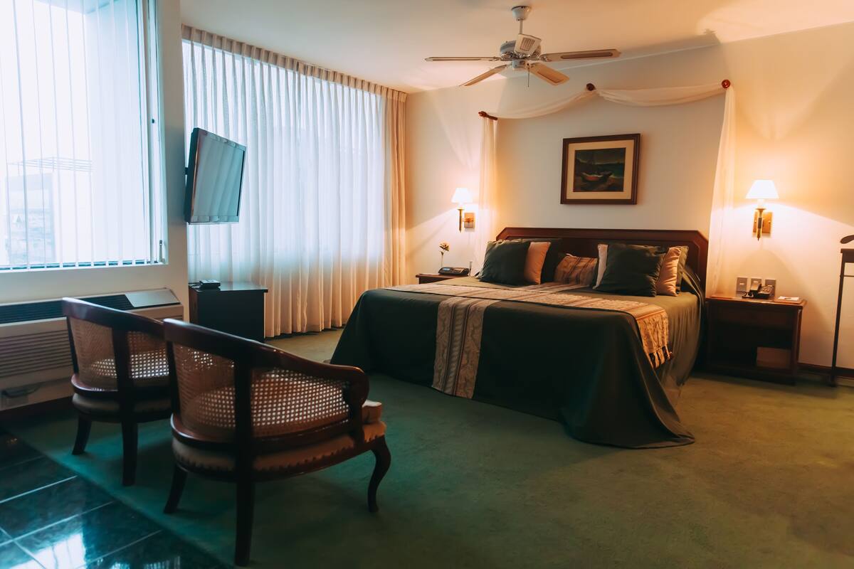 A hotel room with a bed, two bedside tables, a ceiling fan, a TV mounted on the wall, and a window with curtains.