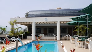 Un área de piscina con sillones de descanso, un espacio de estar techado y un edificio con fachada de vidrio.