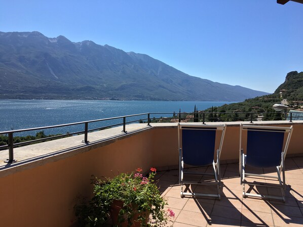 A balcony with two blue chairs, a potted plant with pink flowers, and a view of a lake with mountains in the background. - Villa Belvedere Hotel (Limone sul Garda)