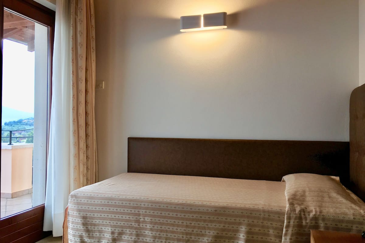 Een slaapkamer met een bed, een nachtkastje, een wandlamp en uitzicht op het groen door een raam met gordijnen.