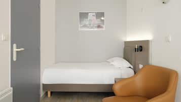 Een eenpersoonsbed met een witte matras en een bruin hoofdbord, een leren fauteuil in bruin, en een wandlamp.