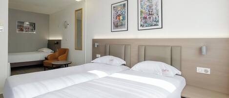 A hotel room with two beds, a chair, a small table, and two framed pictures on the wall.