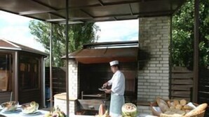 Un'area cucina esterna con uno chef che prepara il cibo, un bancone in mattoni con diversi alimenti e un muro di mattoni sullo sfondo.