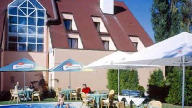 Una piscina con lettini e tavoli, circondata da un edificio con un tetto particolare e ampie finestre.