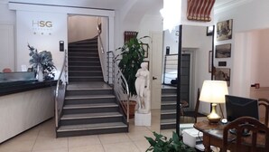 A hotel lobby with a reception desk, a staircase, a statue, and a dining area with a lamp and chair. - Hotel San Giorgio (Bergamo)