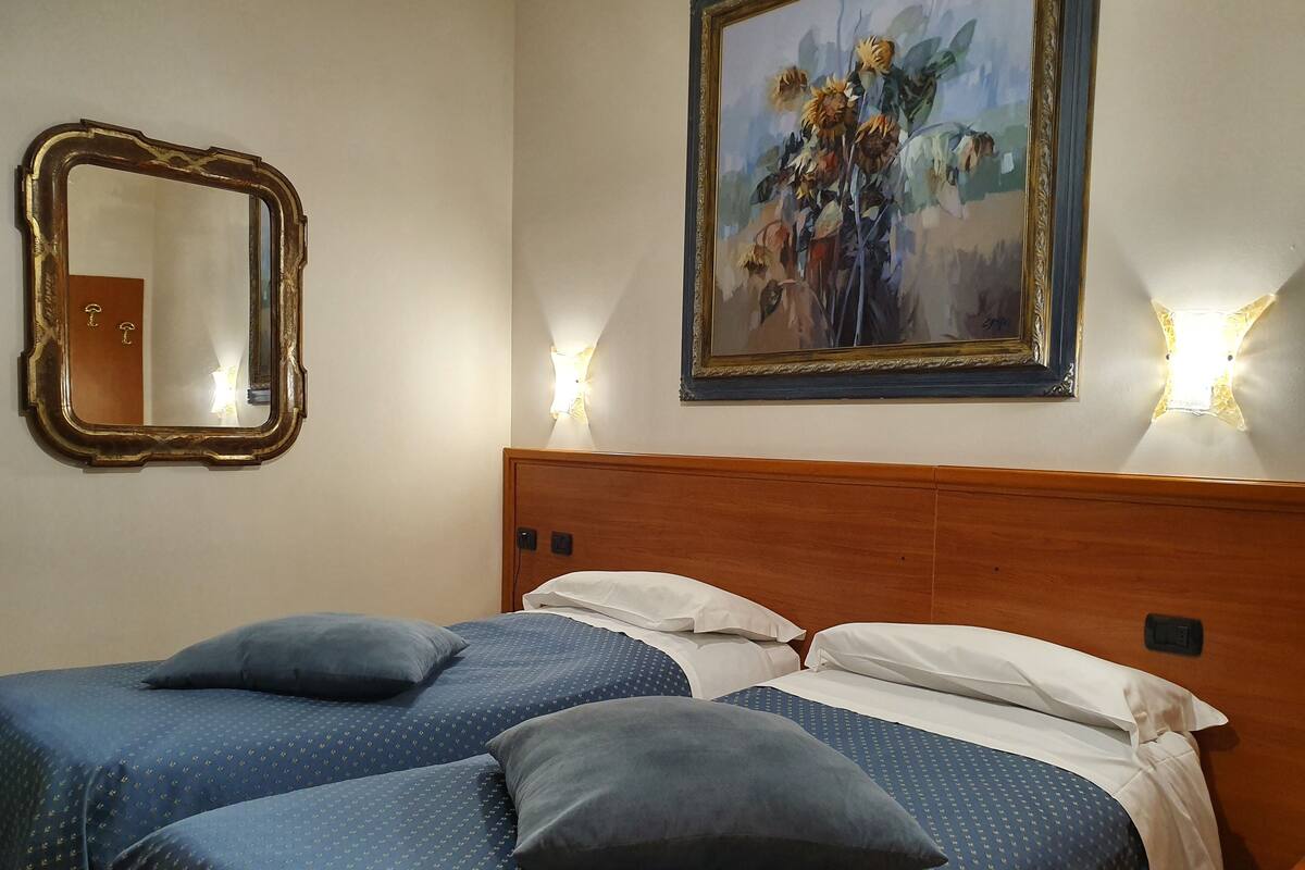 Une chambre d’hôtel avec deux lits, une tête de lit en bois, un tableau représentant des tournesols, un miroir et des lampes fixées au mur.