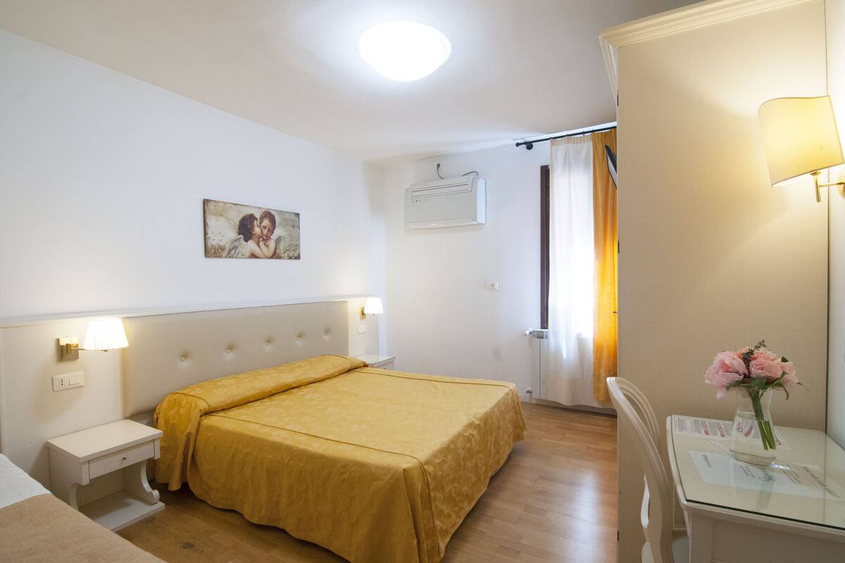 A hotel room with a double bed, a bedside table, a lamp, a painting on the wall, and a vase with flowers on a table.