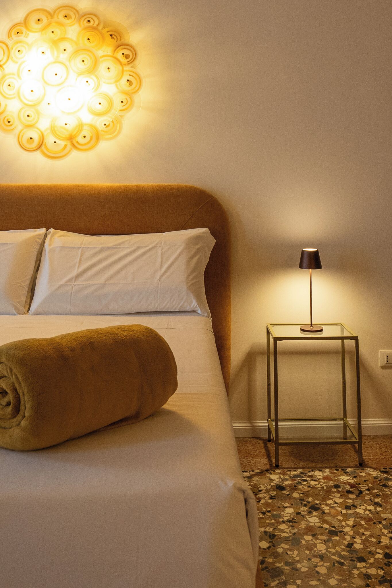A neatly made bed with white linens, a rolled-up towel, a wall-mounted light fixture, and a small table with a lamp beside the bed.