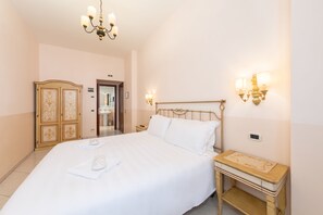 Hotellihuone, jossa on suuri sänky, puinen yöpöytä, pieni pöytä, vaatekaappi ja seinävalaisimet.