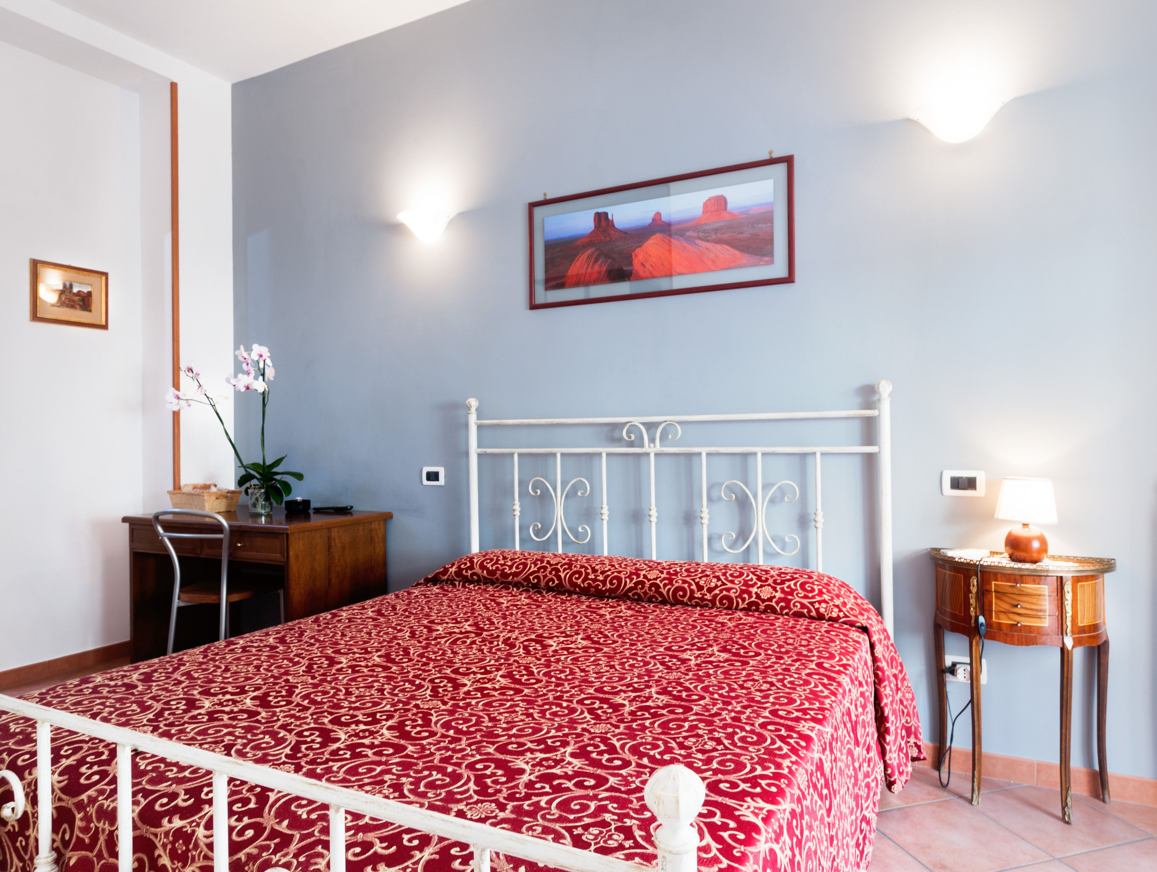 Quarto com cama, mesa de cabeceira, abajur, quadro na parede e escrivaninha com planta.