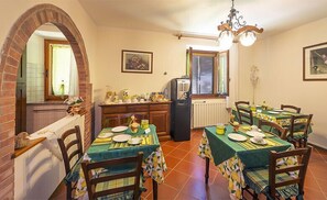 A dining area with six tables, each set with plates, cutlery, and a green tablecloth. There is a brick archway leading to a window, a radiator, and a refrigerator in the background. - Agriturismo Il Colombaiolo (Pienza)