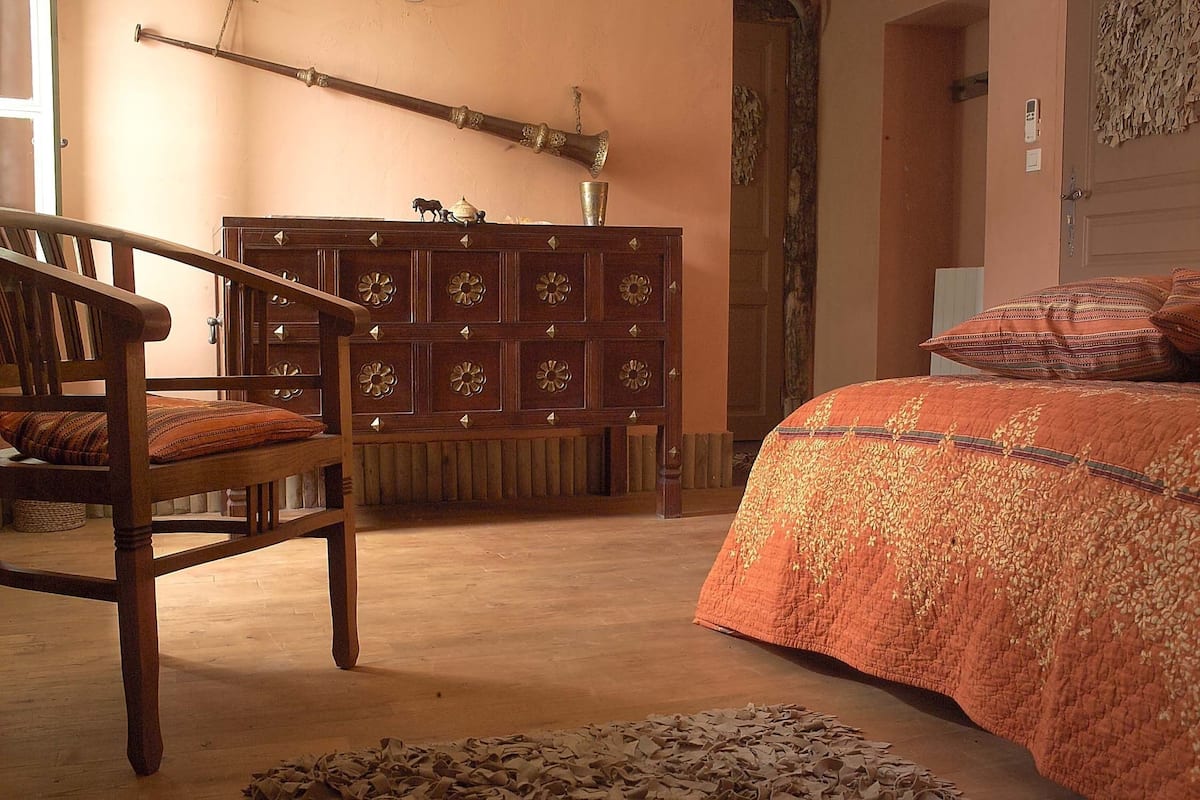 A bedroom with a wooden chair, a bed with an orange quilt, and a wooden cabinet with decorative panels.