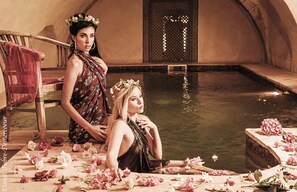 Two individuals seated by a pool with water lilies and candles, in a room with a wooden bench and a door with lattice design.