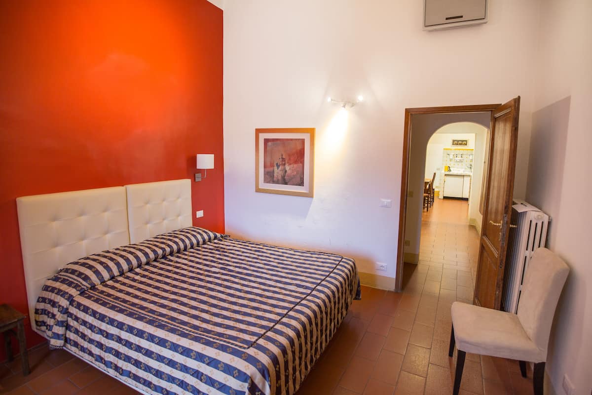 Un dormitorio con cama, una silla, un cuadro y un pasillo que conduce a una cocina.