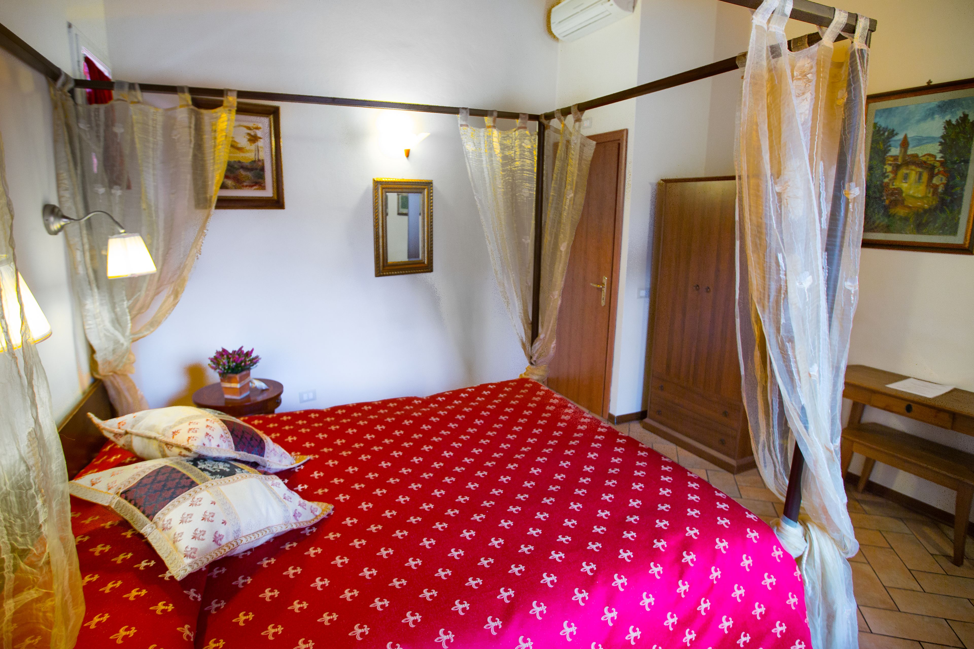 A bedroom with a red bedspread, a wooden wardrobe, a bedside table with a lamp, and a painting on the wall.