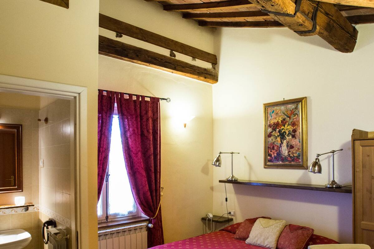 Un dormitorio con cama, mesita de noche, escritorio, silla, ventana con cortinas, radiador y techo de madera.