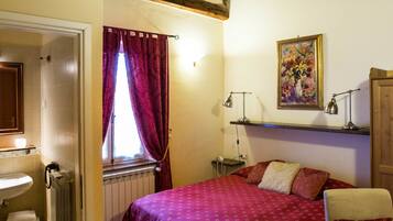 A bedroom with a bed, a nightstand, a desk, a chair, a window with curtains, a radiator, and a wooden ceiling.