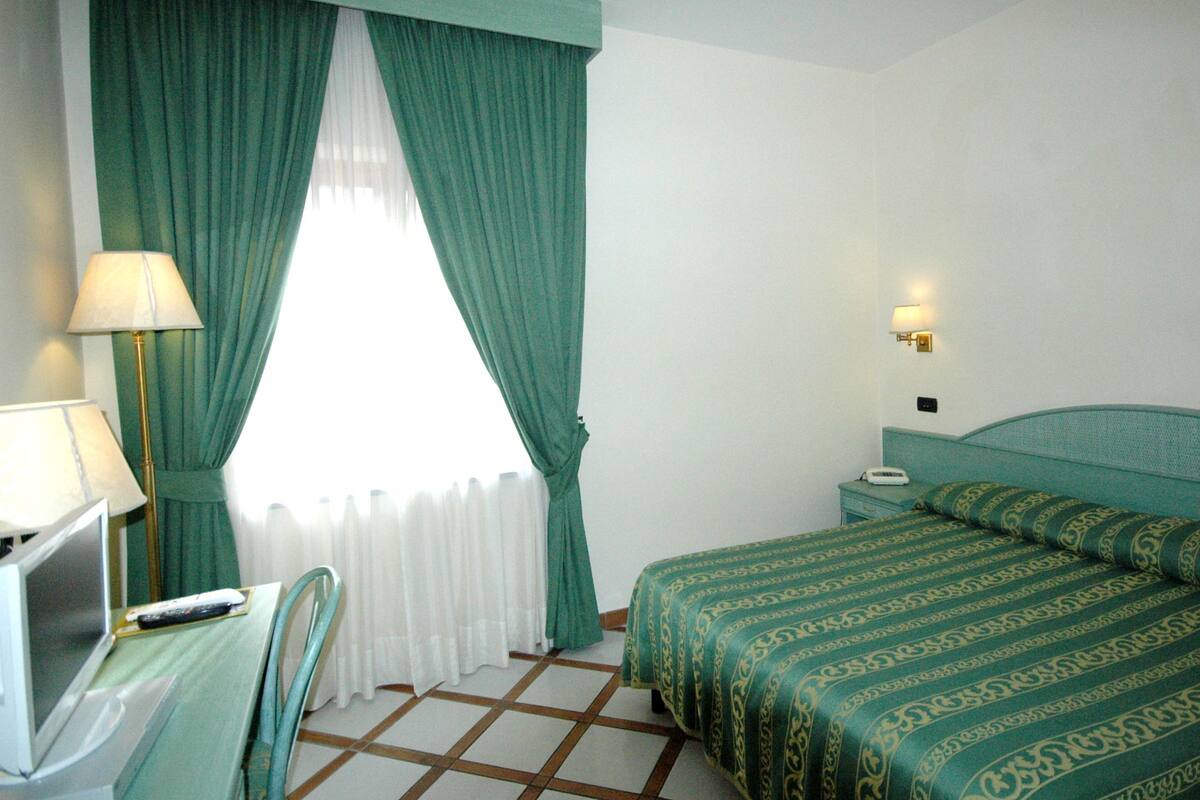 A hotel room with a bed, a desk with a lamp, a chair, and a nightstand with a phone.