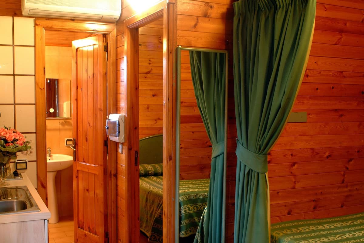 A wooden cabin interior with a bathroom, a sink, a bed with green curtains, and a small kitchenette.