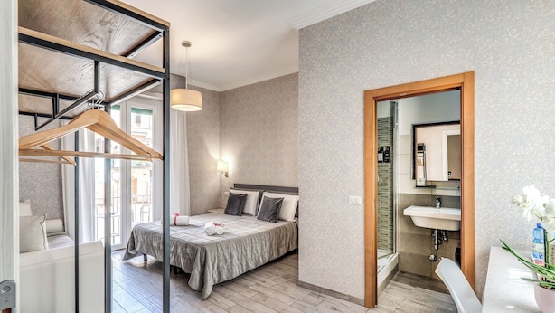 A bedroom with a bed, bedside table, hanging clothes rack, and a bathroom with a sink and mirror. - Marta Inn (Rome)