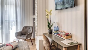 A modern hotel room with a bed, a grey armchair, a small desk with a plant, a TV on the wall, and a window with sheer curtains. - Marta Inn (Rome)