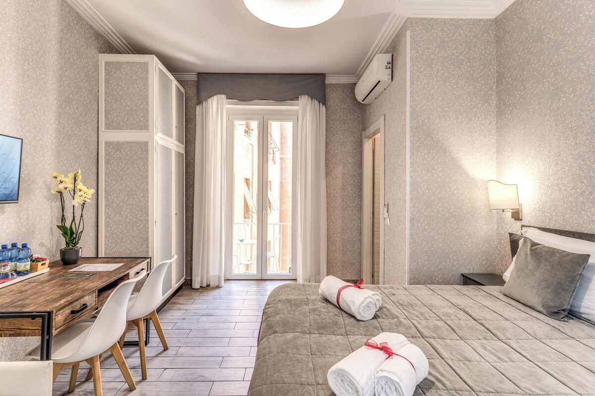 A hotel room with a bed, a desk with chairs, a window with curtains, and a wall-mounted air conditioner.